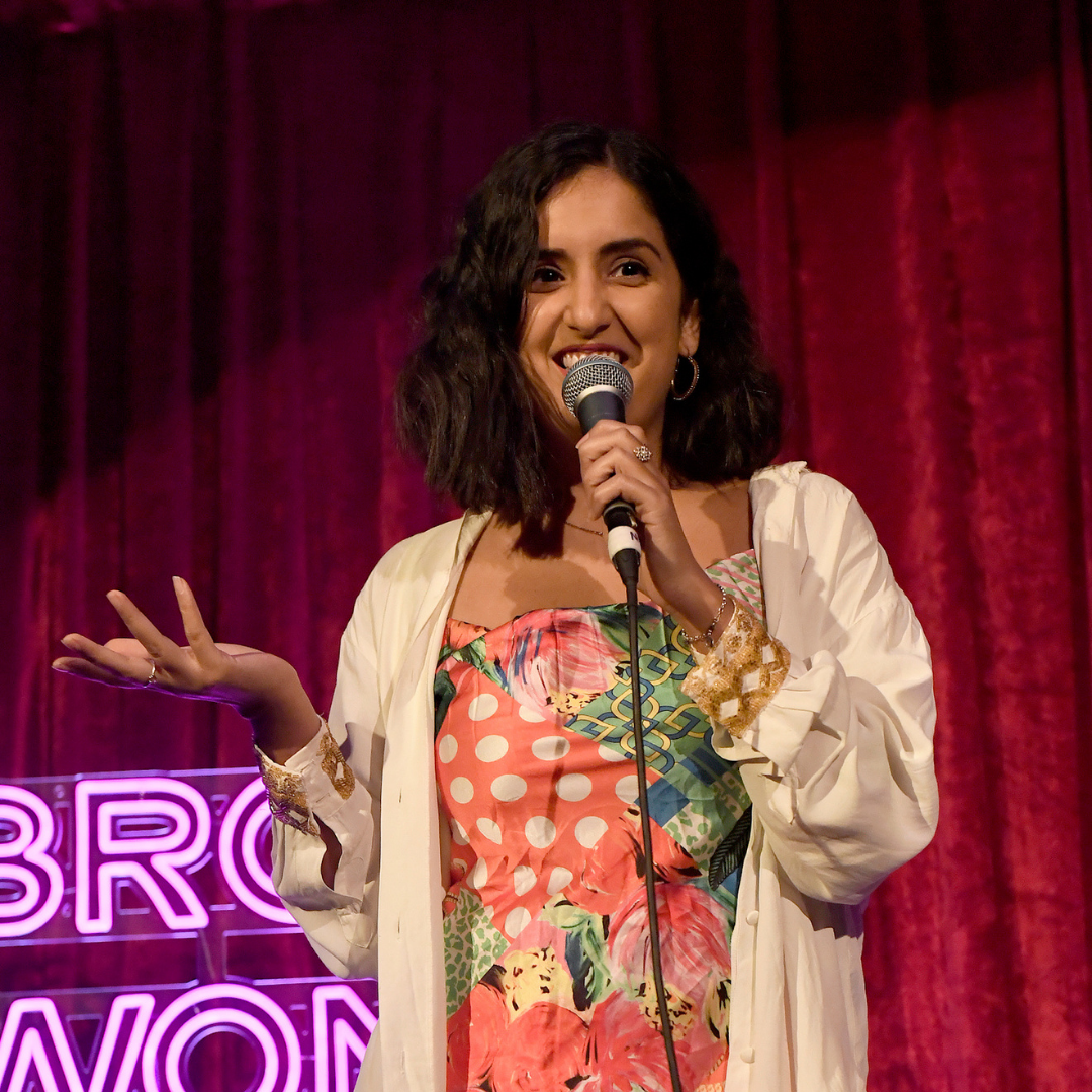 A woman performs stand-up comedy on stage, smiling with a microphone in front of a red curtain and neon sign.